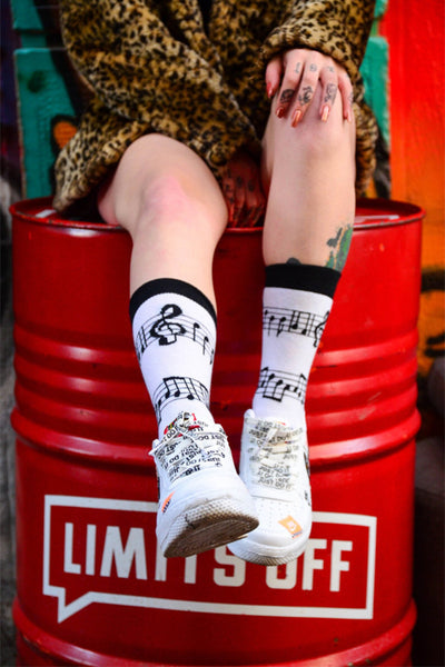 A person seated on a red barrel with legs crossed, showcasing white sneakers and socks with a black musical note pattern.