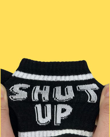stretching SHUT UP written black colour socks from its ankle part on yellow background
